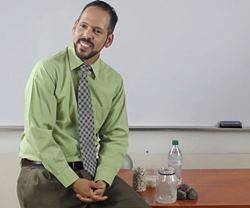 Un profesor apoyado en su mesa con un frasco al lado.