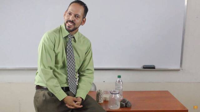 Un profesor apoyado en su mesa con un frasco al lado.